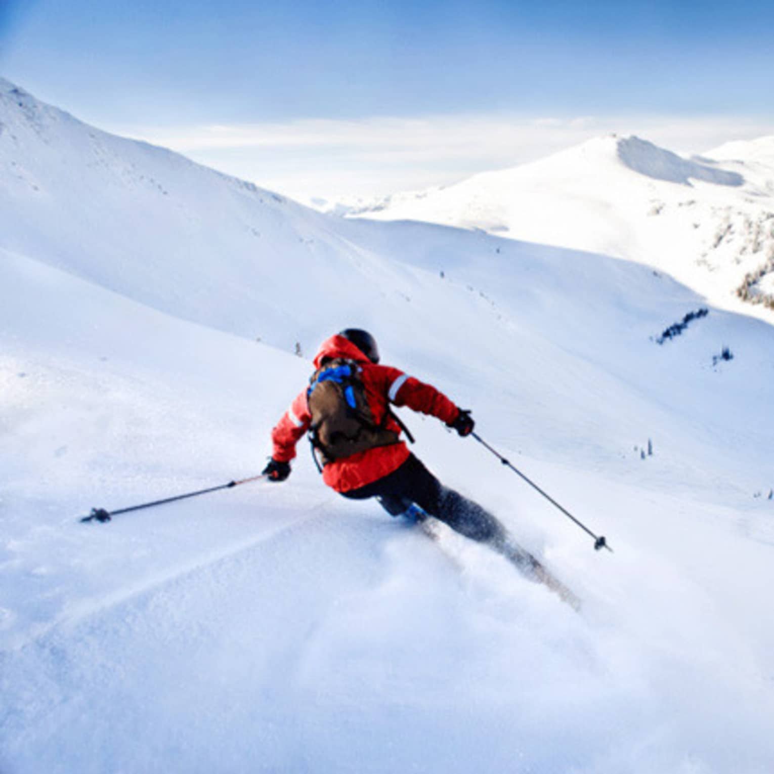 Back view of skier in red jacket, backpack, skiing down mountain slope on sunny day
