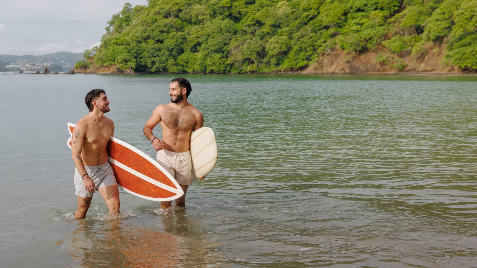 Two people walk out of the water, each holding a surfboard under their arm