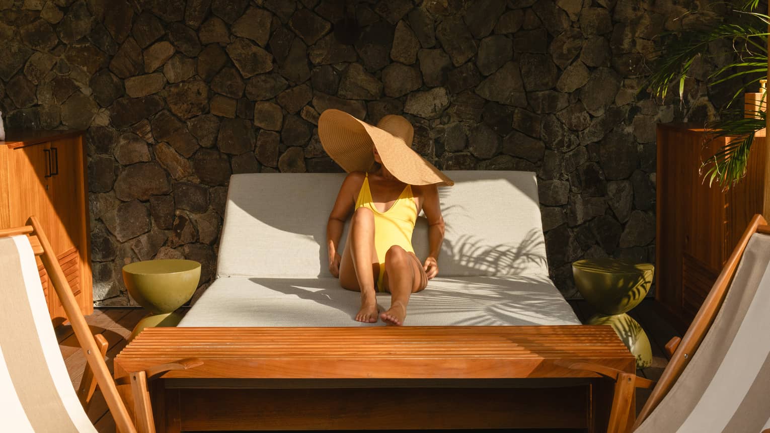 Guest wearing a yellow bathing suit and a large straw sunhat lounges on a poolside daybed