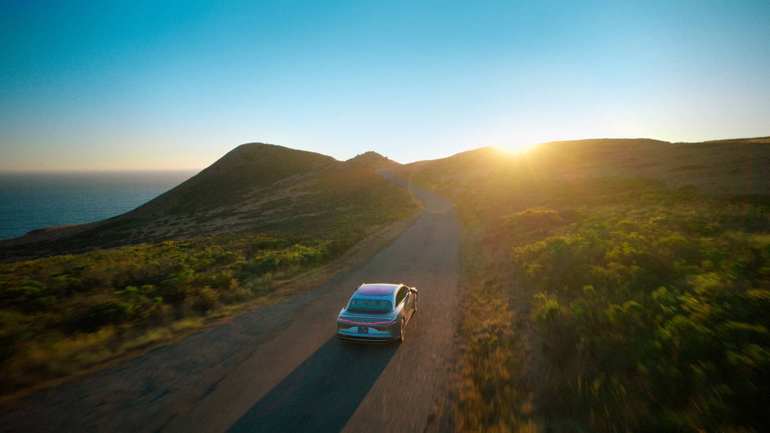 A car drives down a road surrounded by green hilly terrain. A clear blue sky with the sun just peeking over the hills is in the background, and there is also a glimpse of the ocean behind the hills.