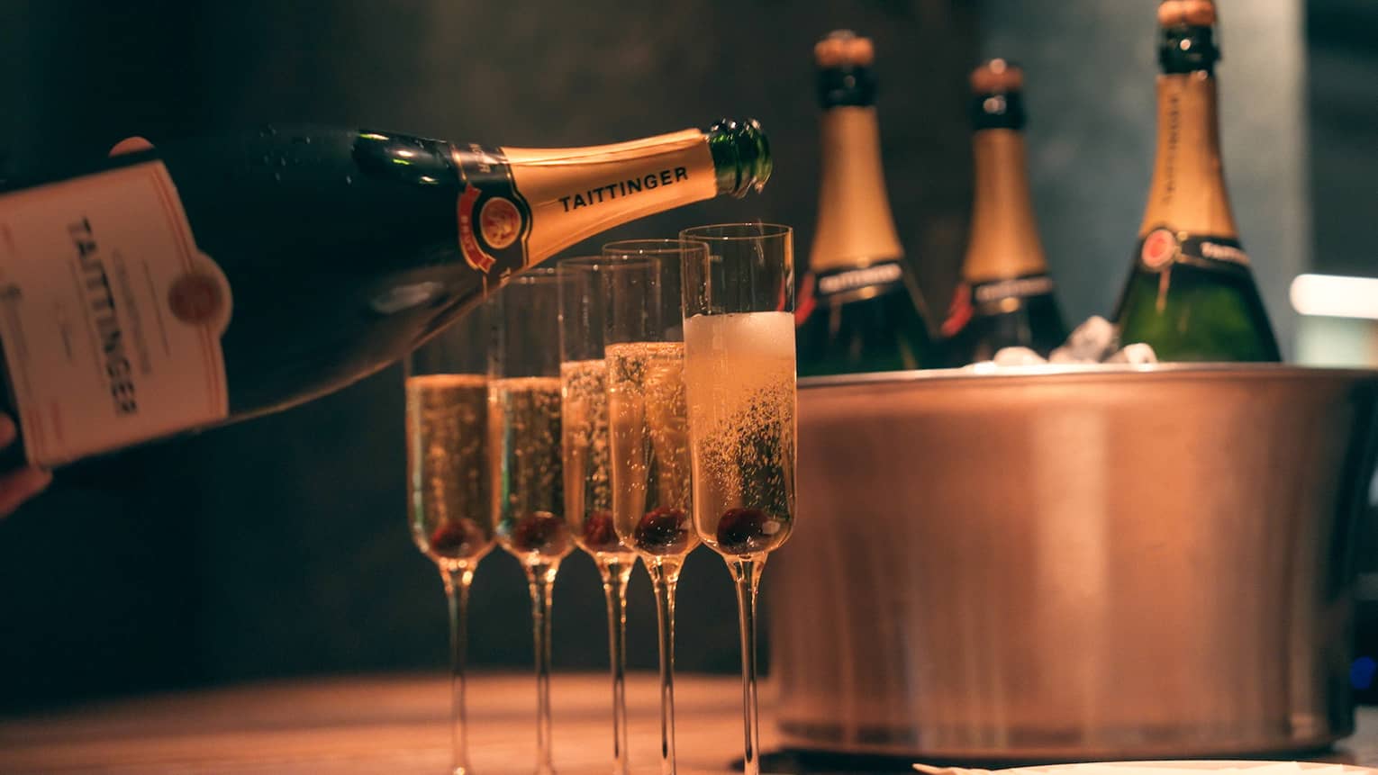 Bottle of champagne being poured into four champagne glasses beside copper ice bucket with three champagne bottles