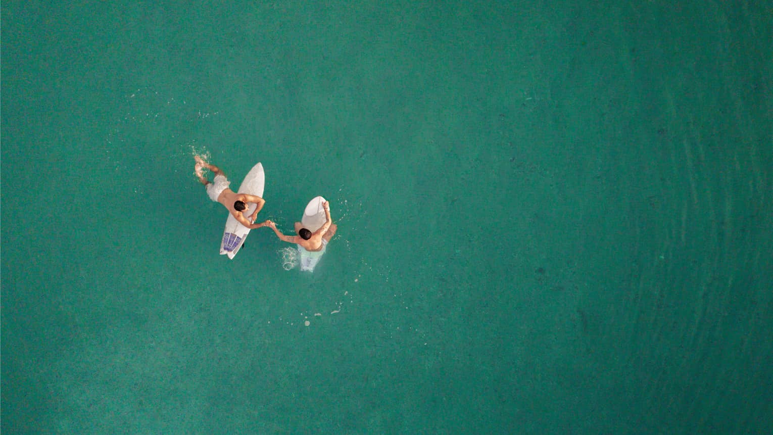 Aerial view of two people in the blue-green ocean water, each sitting on a surfboard and holidng hands