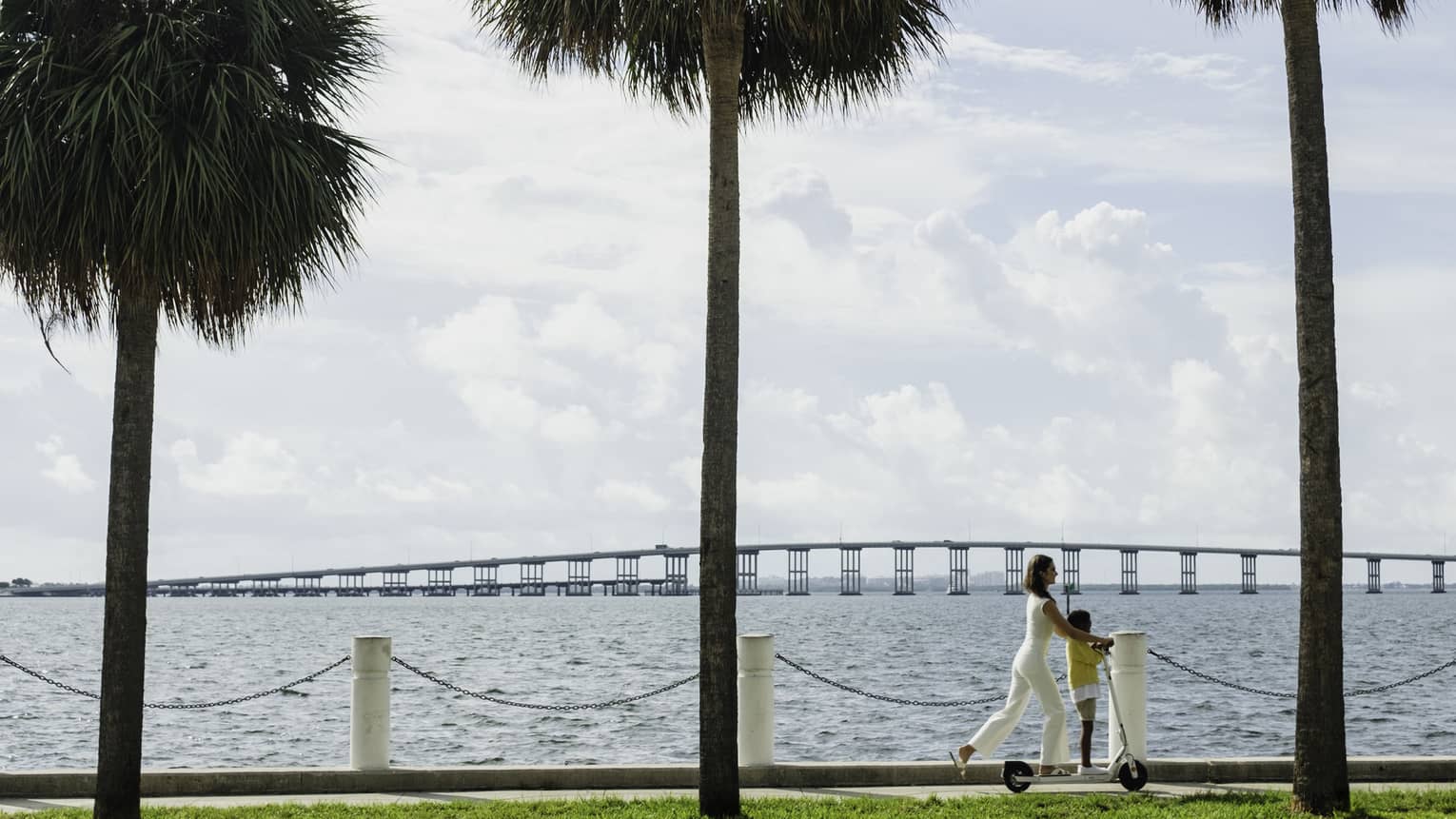 Woman and young boy ride scooter together along the waterway beneath palm trees