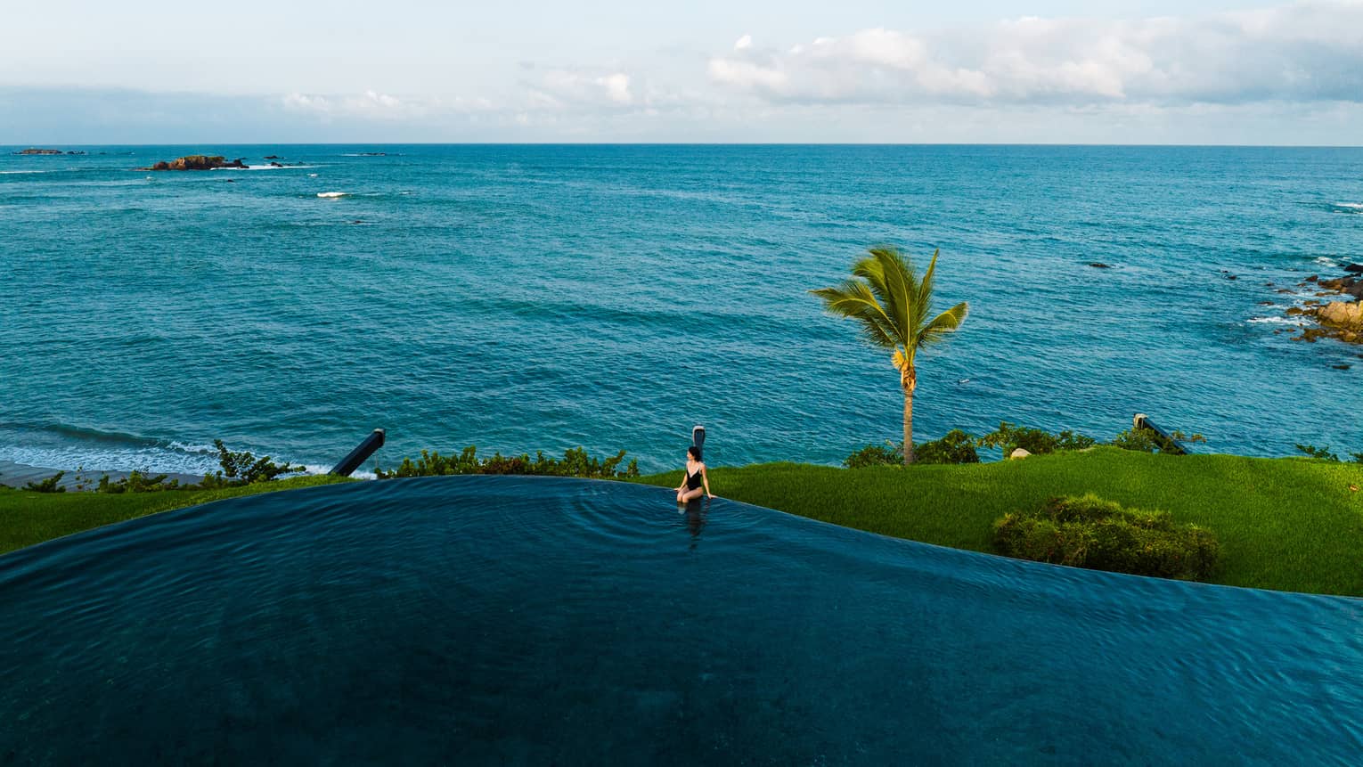 A person sits on the edge of an oceanfront infinity pool at tropical resort