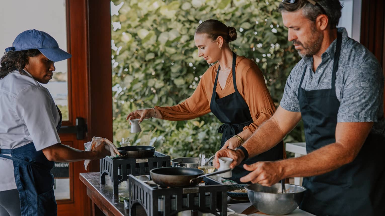 A couple cook a dish with a chef, using portable stoves and kitchenware on a wooden table. The ocean is in the background.