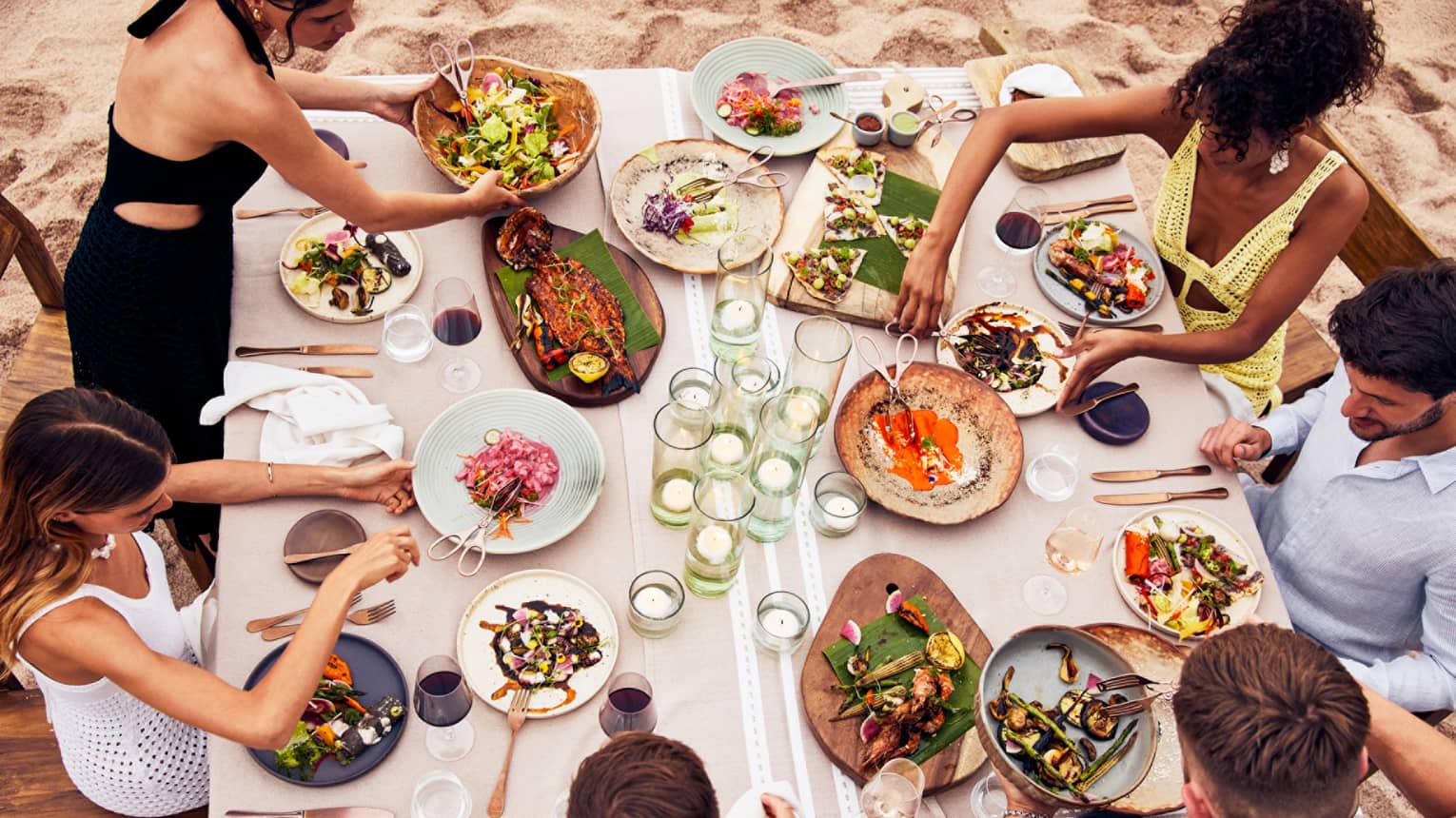 Beachside dining table with an assortment of vibrant dishes, candlelit glass holders and guests enjoying fresh seafood and grilled vegetables