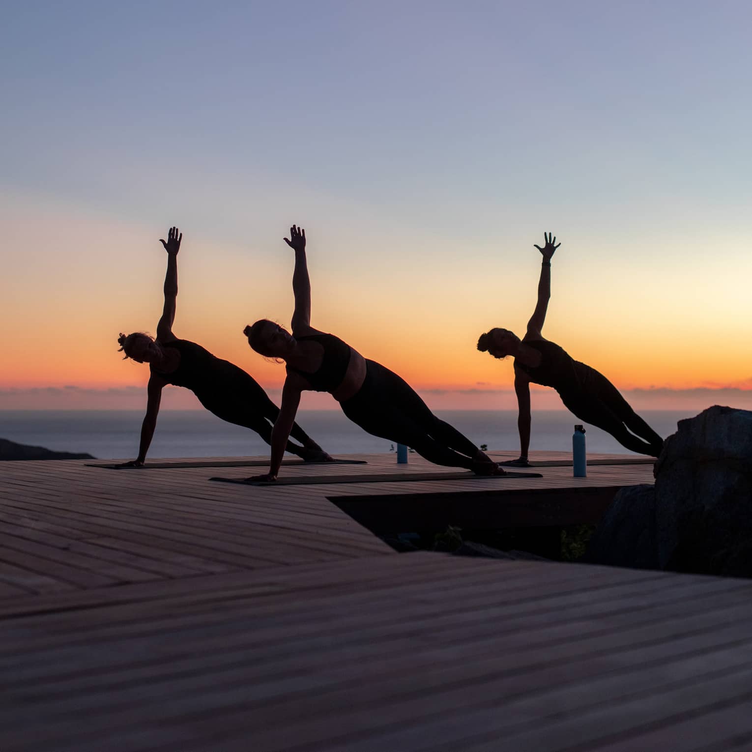 Three guests doing side plank poses on yoga mats on a wooden dock are silhouetted against a clear blue, orange and pink sky.