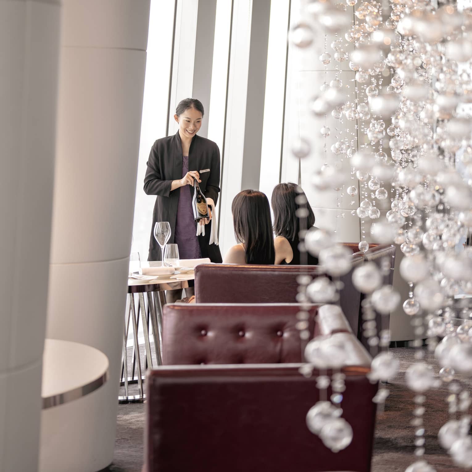 A server presents a bottle of wine to two diners in a leather tufted banquette, a cascade of shimmering white baubles behind.