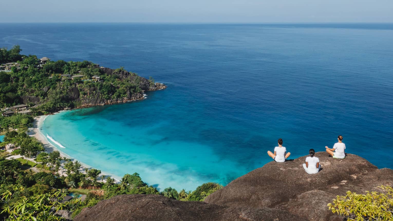 Three people sit cross-legged in meditation overlooking the ocean.