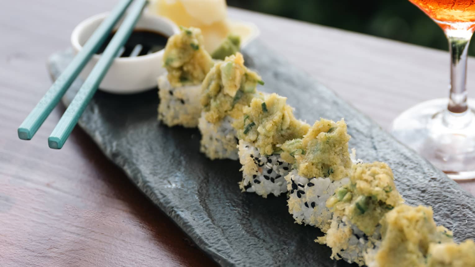 Sushi rolls on tray with chopsticks and wine glass.