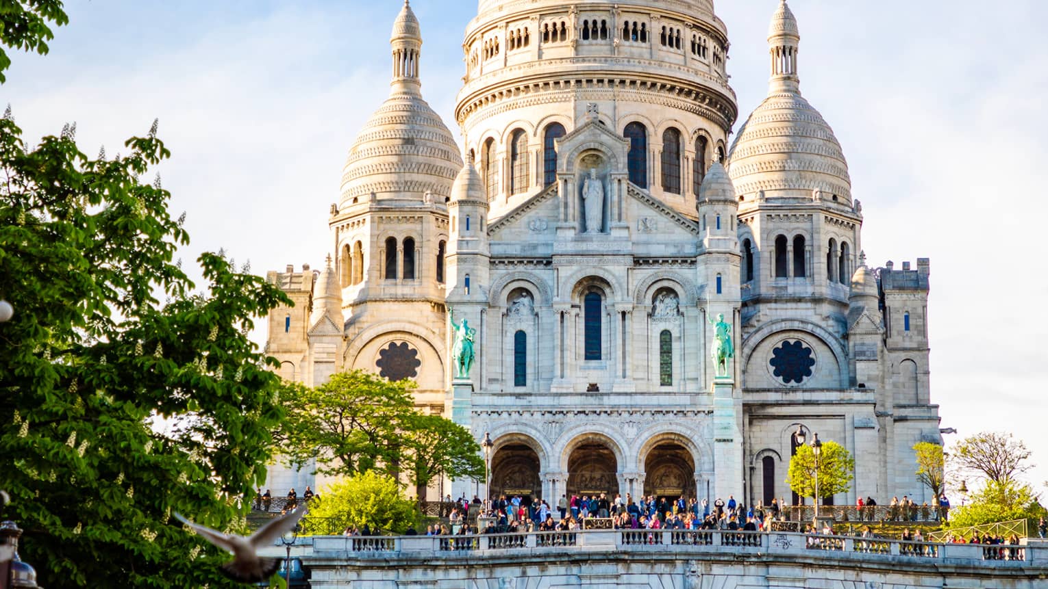 Sacré-Coeur Basilica sits atop a grassy hill under a cloudy blue sky as tourists mill about near the entrance.