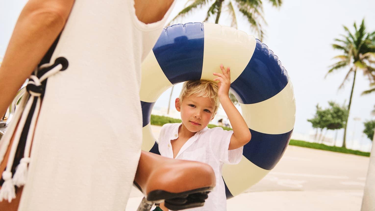 Young child wearing green swim trunks and a white shirt holds a blue-and-cream pool float while walking next to adult wearing a cream-coloured beach cover up and walking next to a bicycle