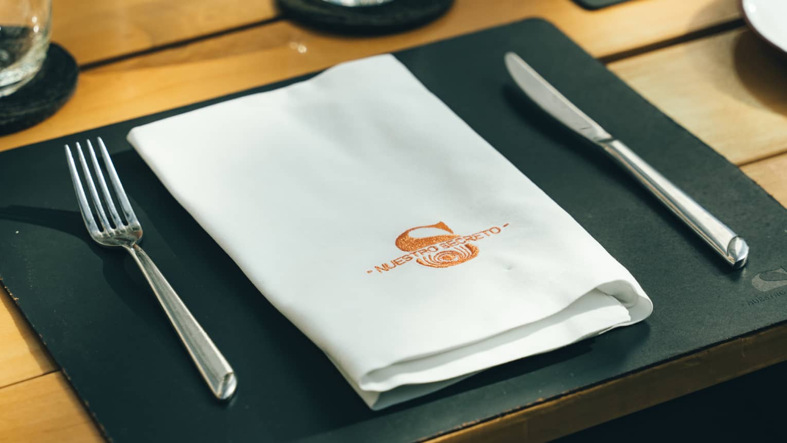 Table setting with a black rectangular placemat, white folded napkin and silver cuttlery on a wood-slat table