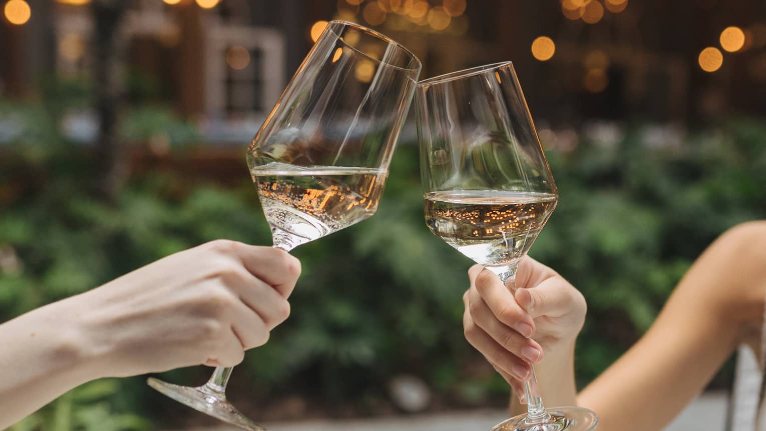 Two guests holding wine glasses, with plates of food on the table and lights in the background.