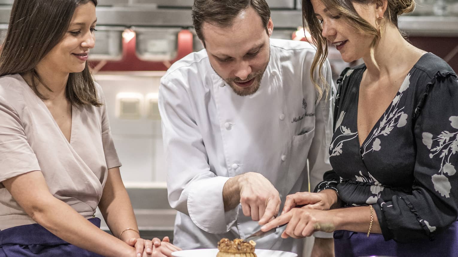 A chef stands between two guests and points to a pastry that one guest is cutting while the other guest smiles and looks on.