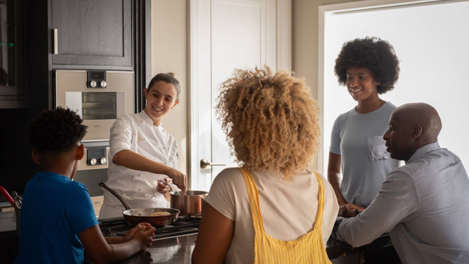 A family of four gathers around a kitchen-island cooktop watching a smiling chef prepare food in copper cookware.