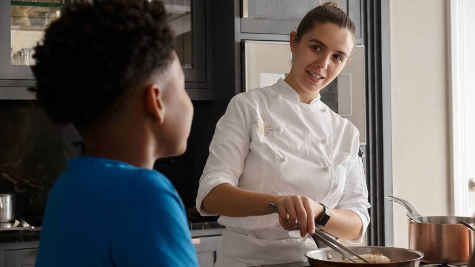 Rear close-up of a child looking on as a smiling chef prepares food in a copper frying pan, a copper pot on a nearby burner.