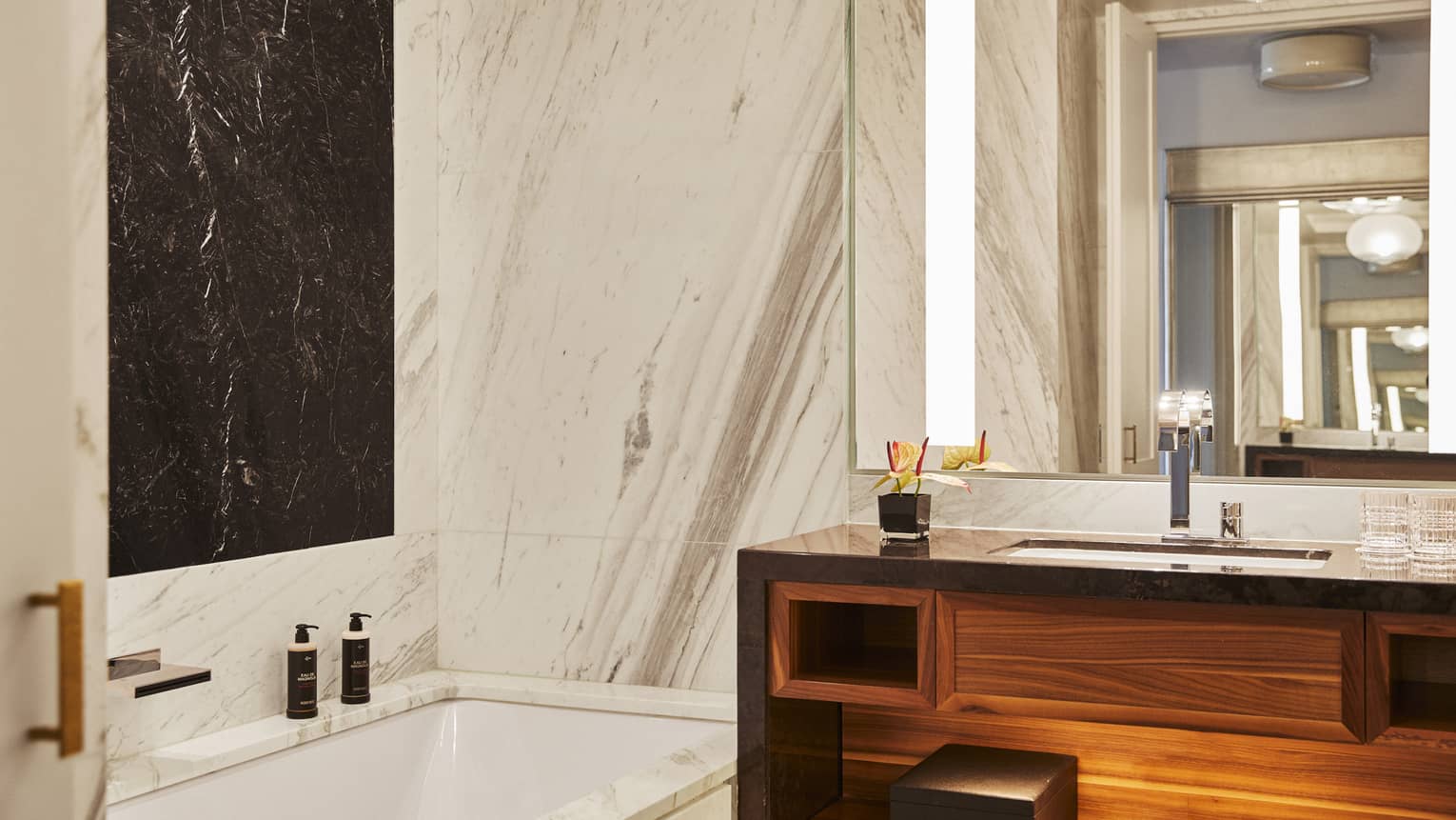 Luxury hotel bathroom with marble walls, black accent panel, built-in bathtub, wooden vanity and large mirror with lighting
