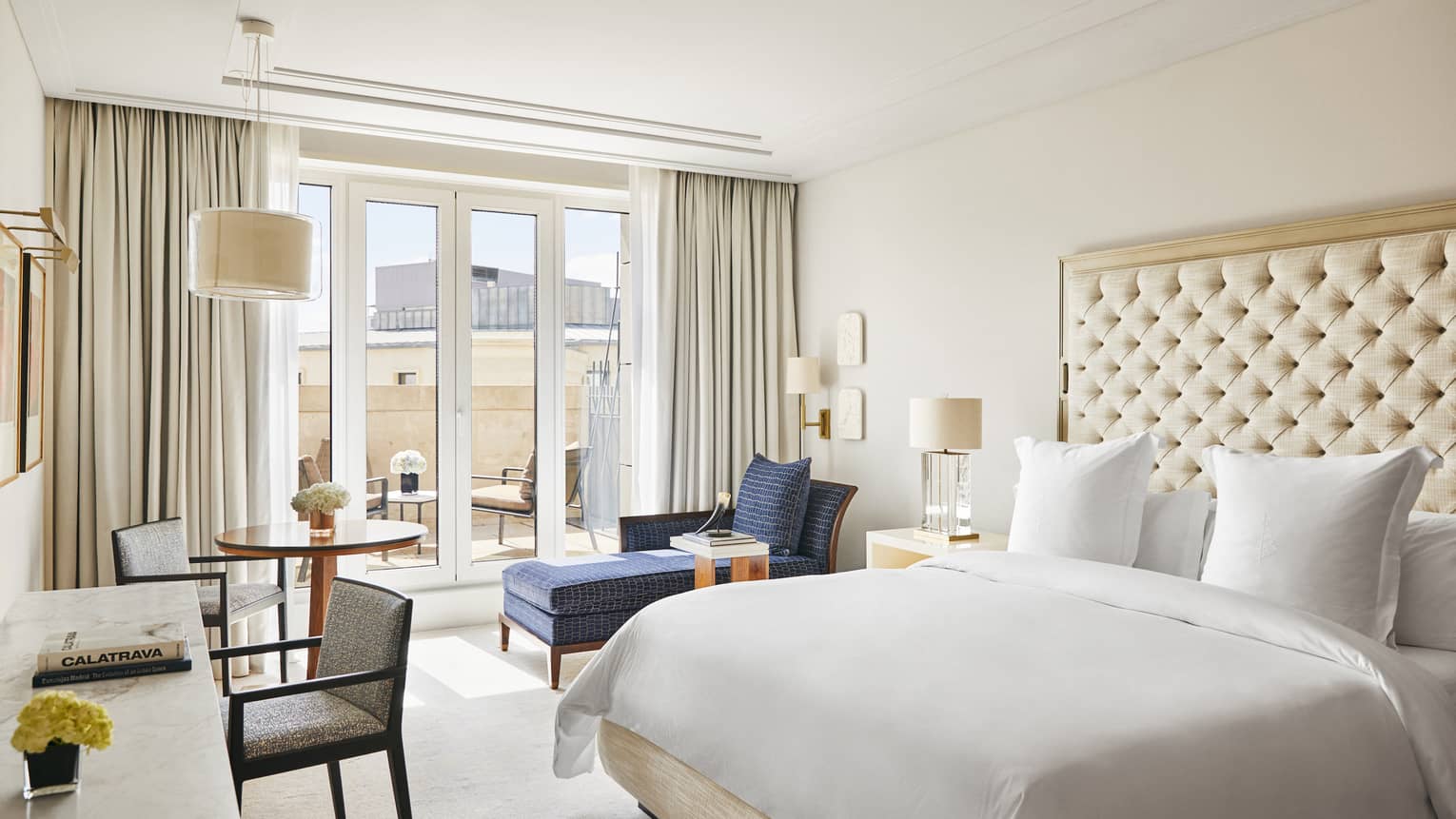Luxury hotel bedroom with a king bed, tufted headboard, blue chaise lounge, round table and desk with chairs and French doors leading to a terrace