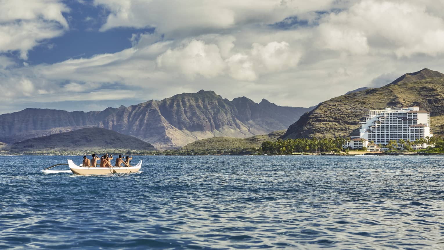 Group of men paddle white canoe on ocean, resort and island in background