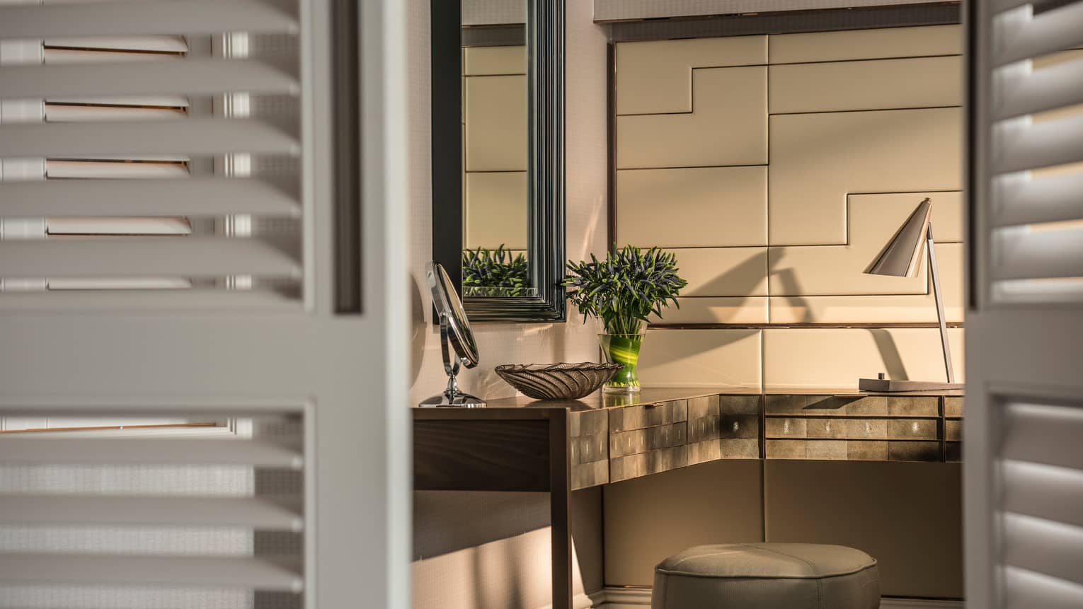 A stylish vanity area with a cushioned stool, sleek desk, decorative mirror and table lamp. A vase of fresh flowers adds a touch of elegance, viewed through shuttered doors.
