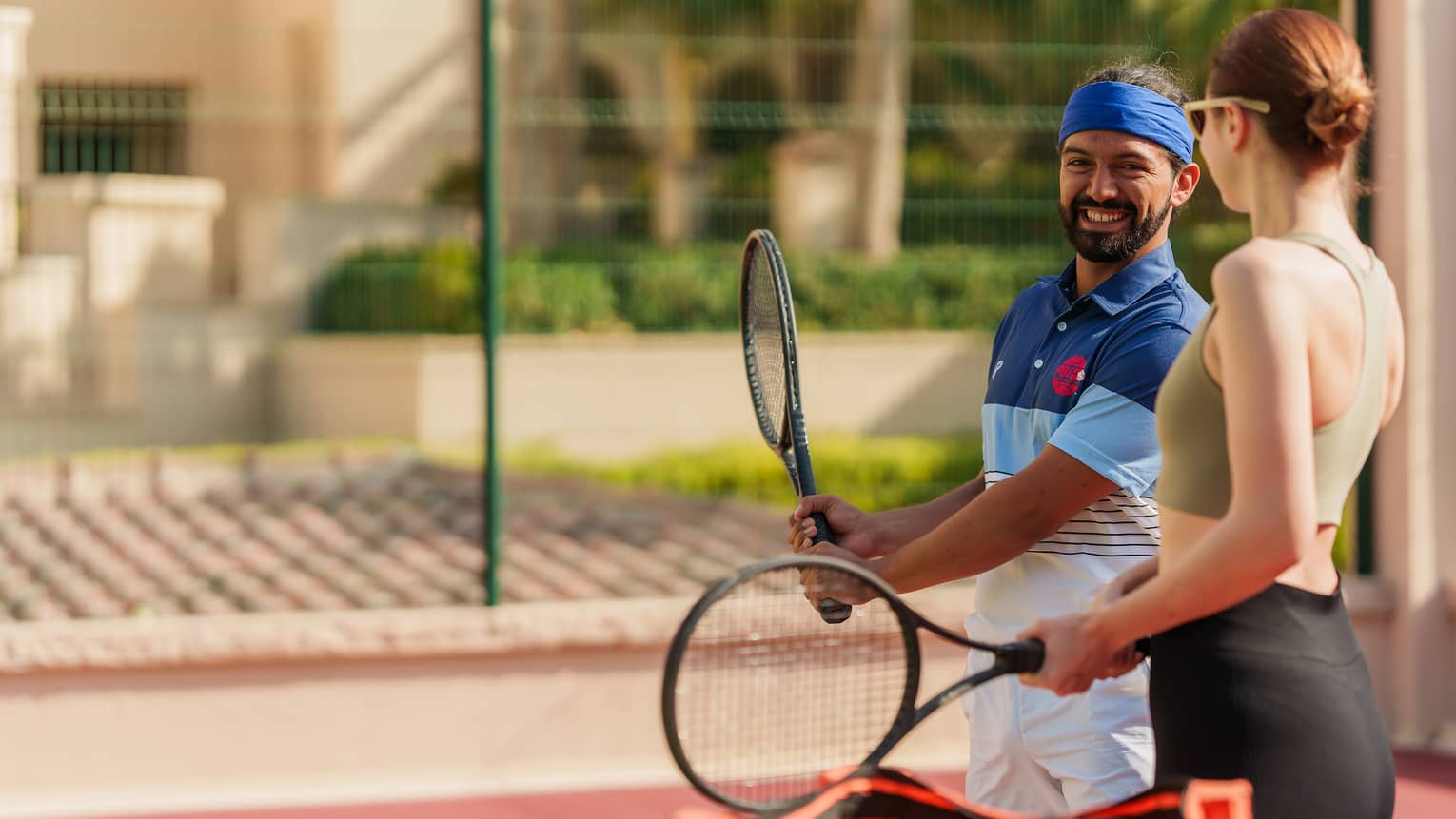 Two guests in athletic wear hold racquets on an outdoor court, one demonstrating a grip to the other, smiling.