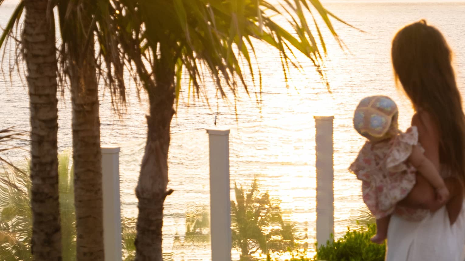 Mother wearing long white dress and holding an infant on her hip walks next to palm trees with the sun rising over the ocean in the distance