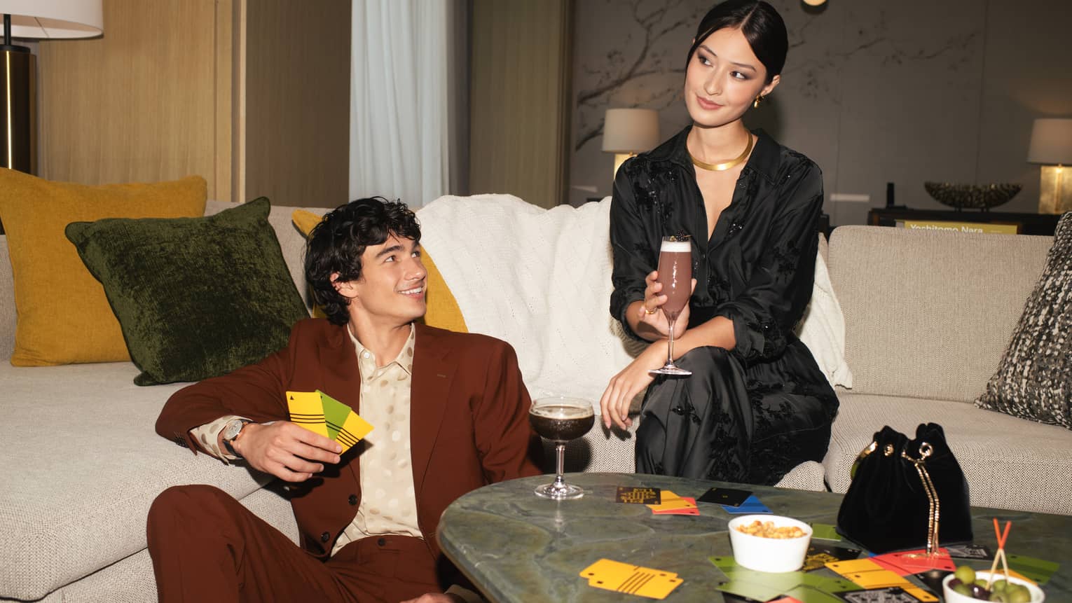 Two people sitting on a sofa with drinks, surrounded by snacks and game cards, enjoying a casual gathering in a stylish living room