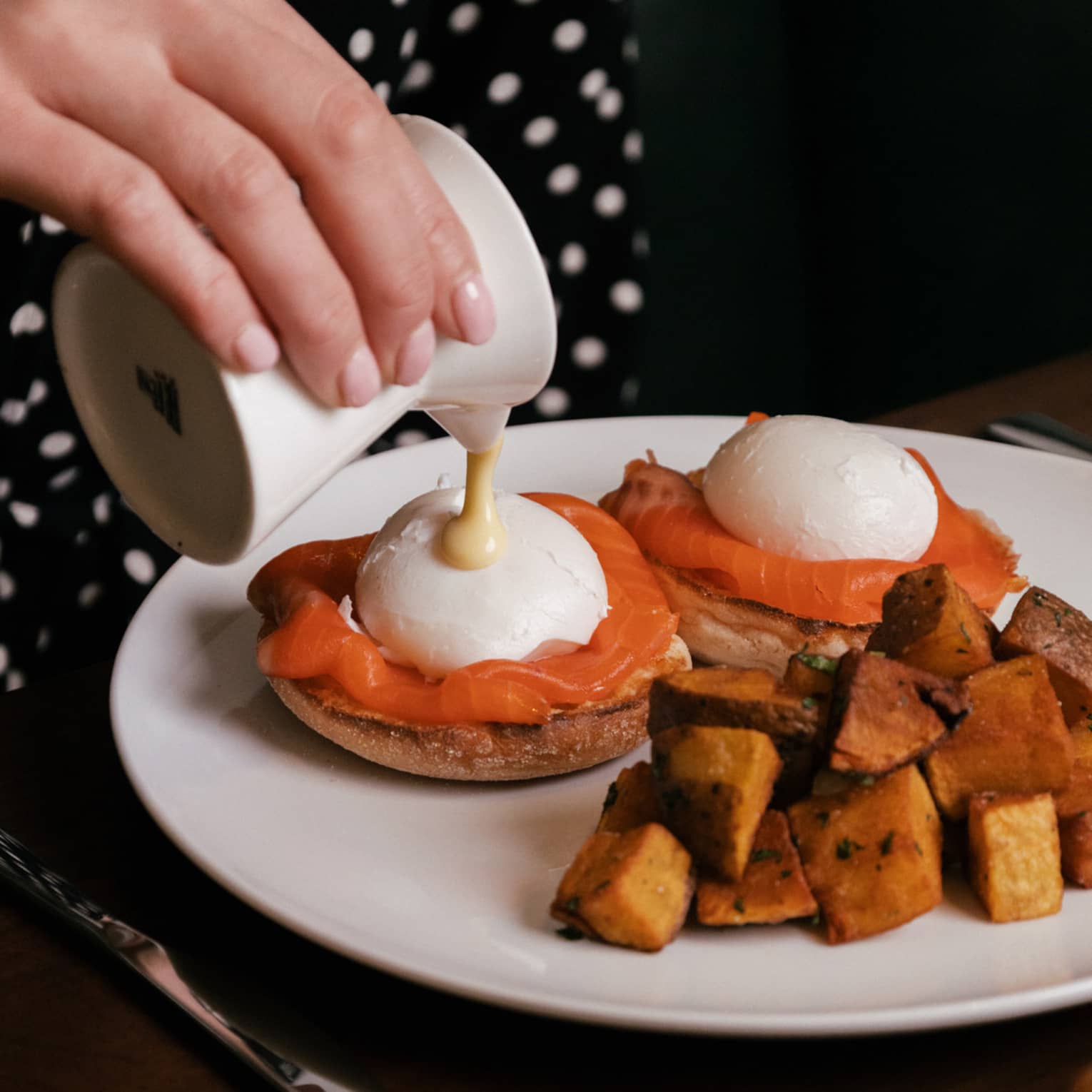 Sauce being poured over two poached eggs with smoked salmon on English muffins, served alongside roasted potatoes.