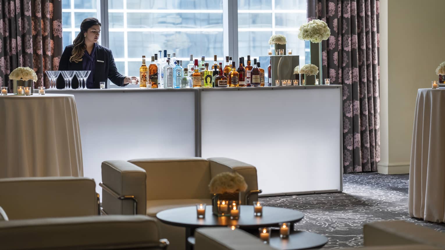 Bartender stands at liquor bottle-lined bar in front of window near seating area