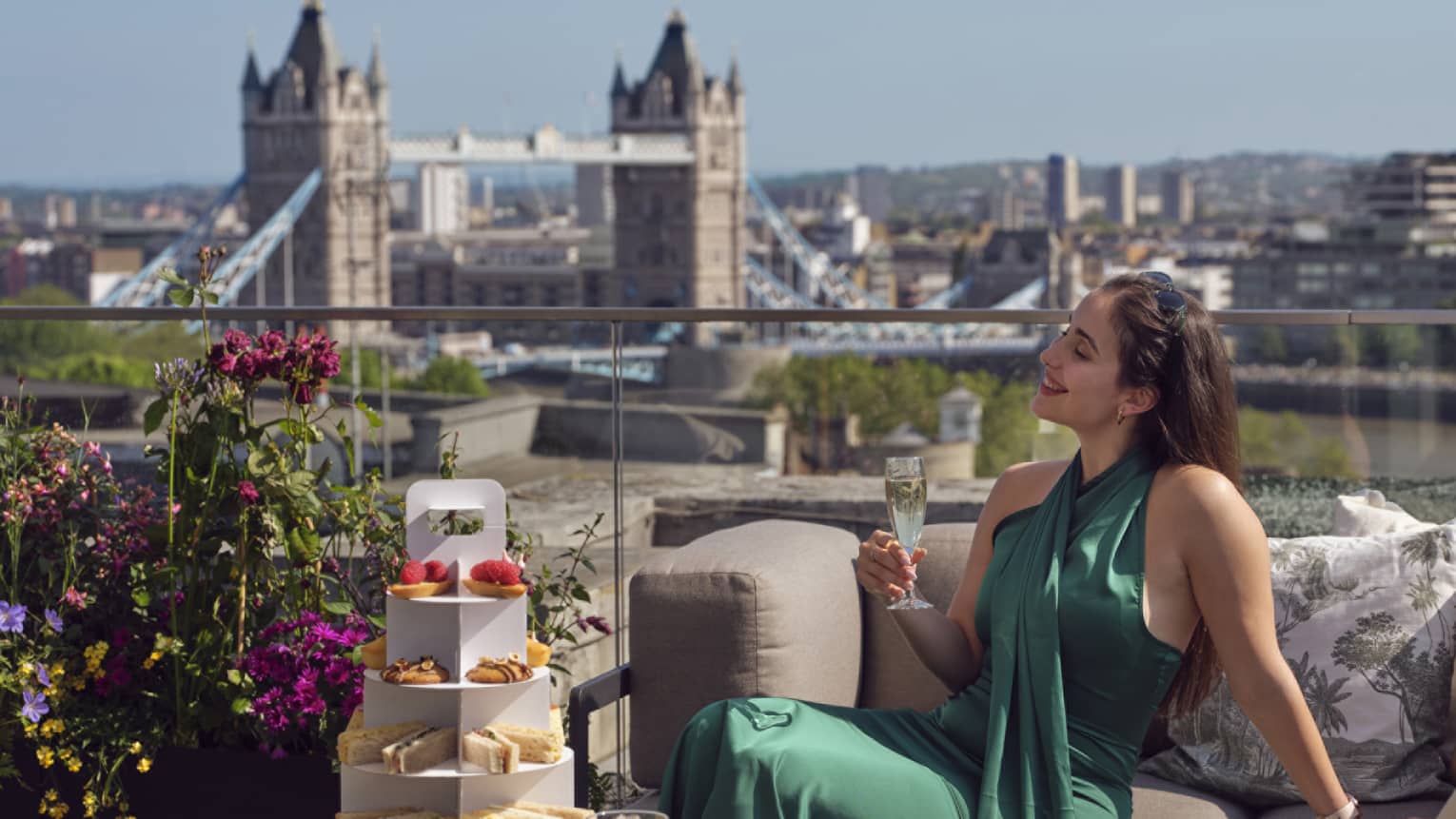 Woman in green dress holding champagne beside tea stand with confections and Tower Bridge in backdrop