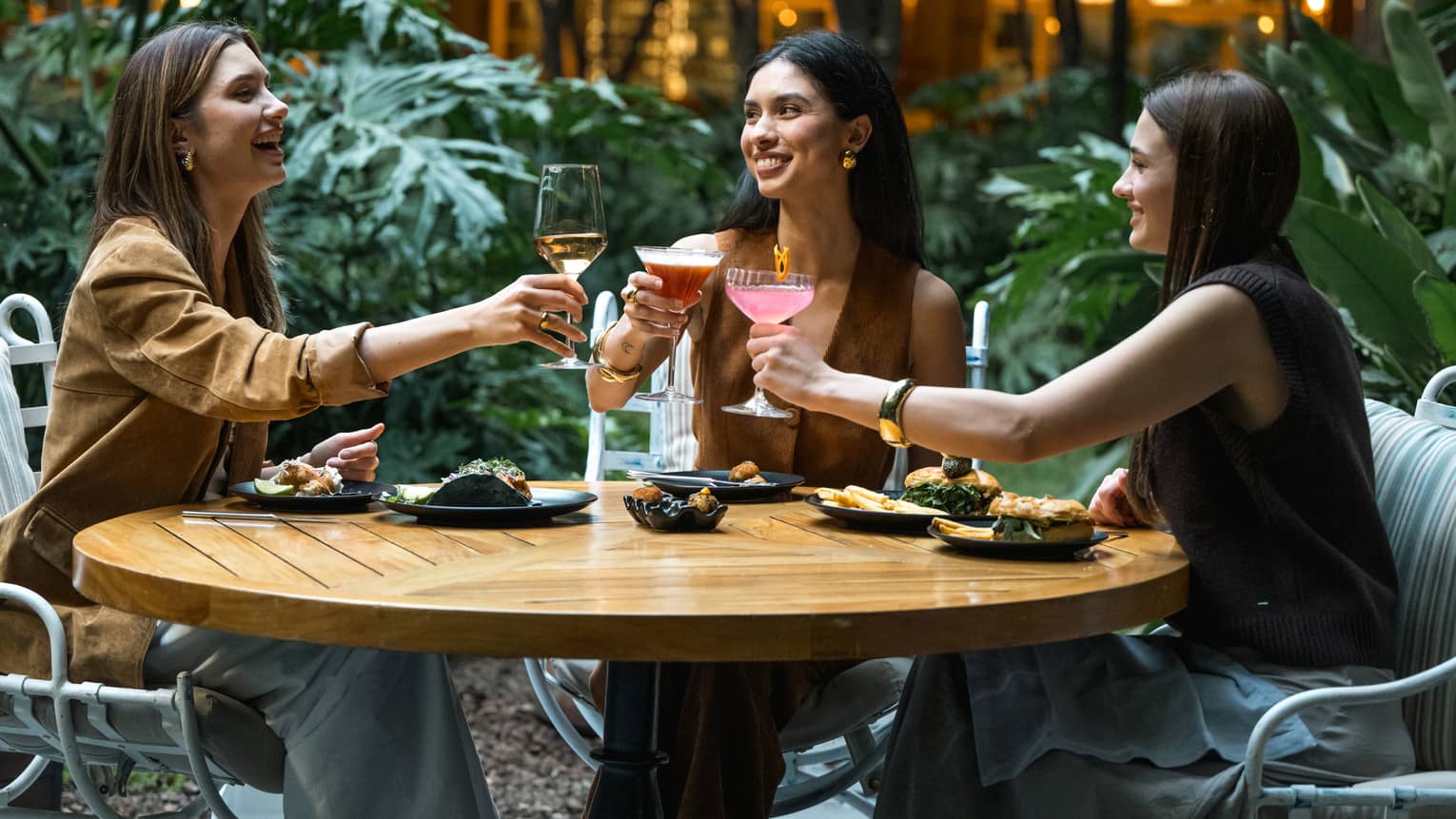 Three guests sit around a round wood table in an outdoor dining space and raise their glasses in a toast.
