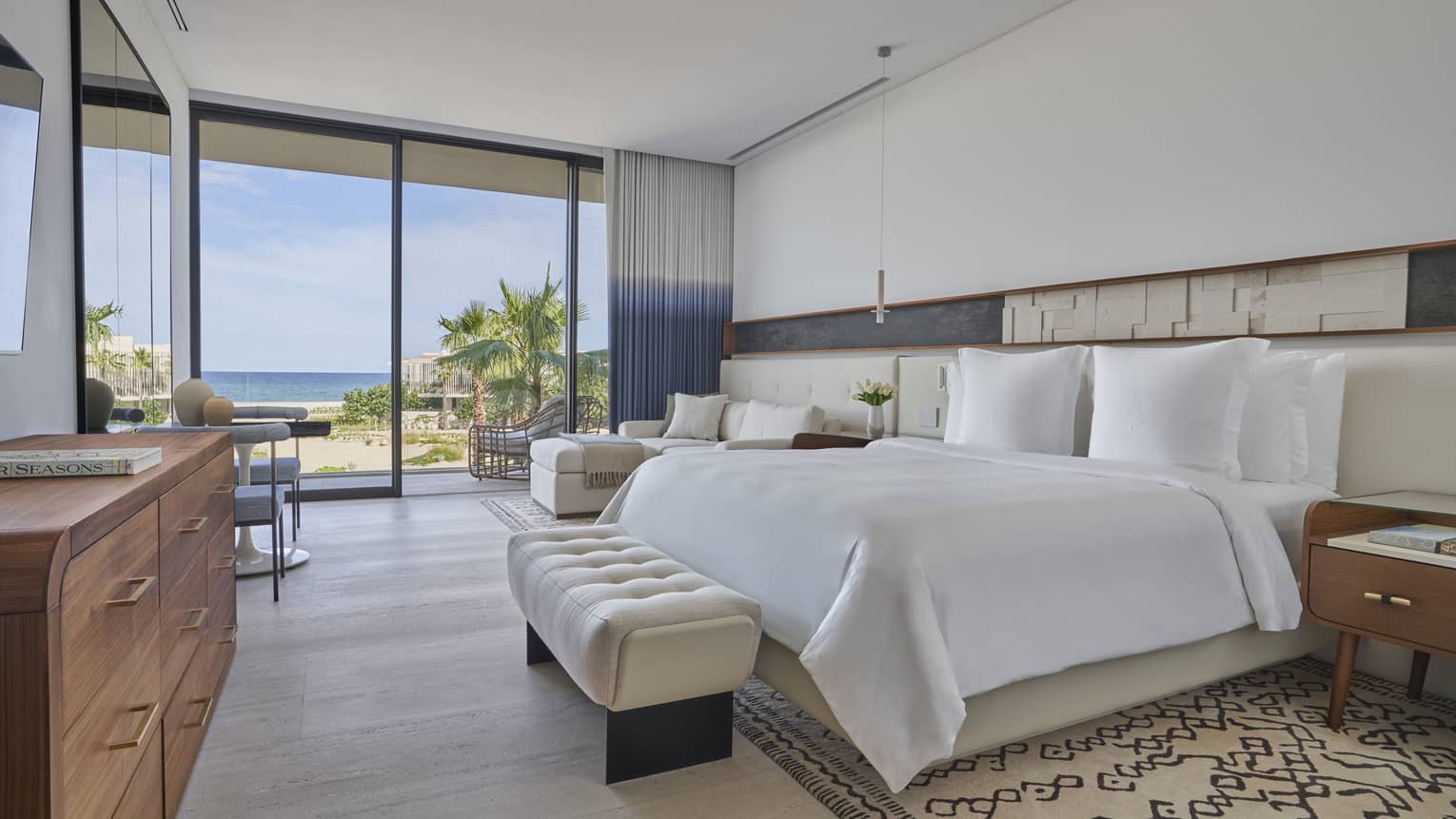 White bed with upholstered foot bench, side tables, dresser, lounge chair, tall glass doors and water views