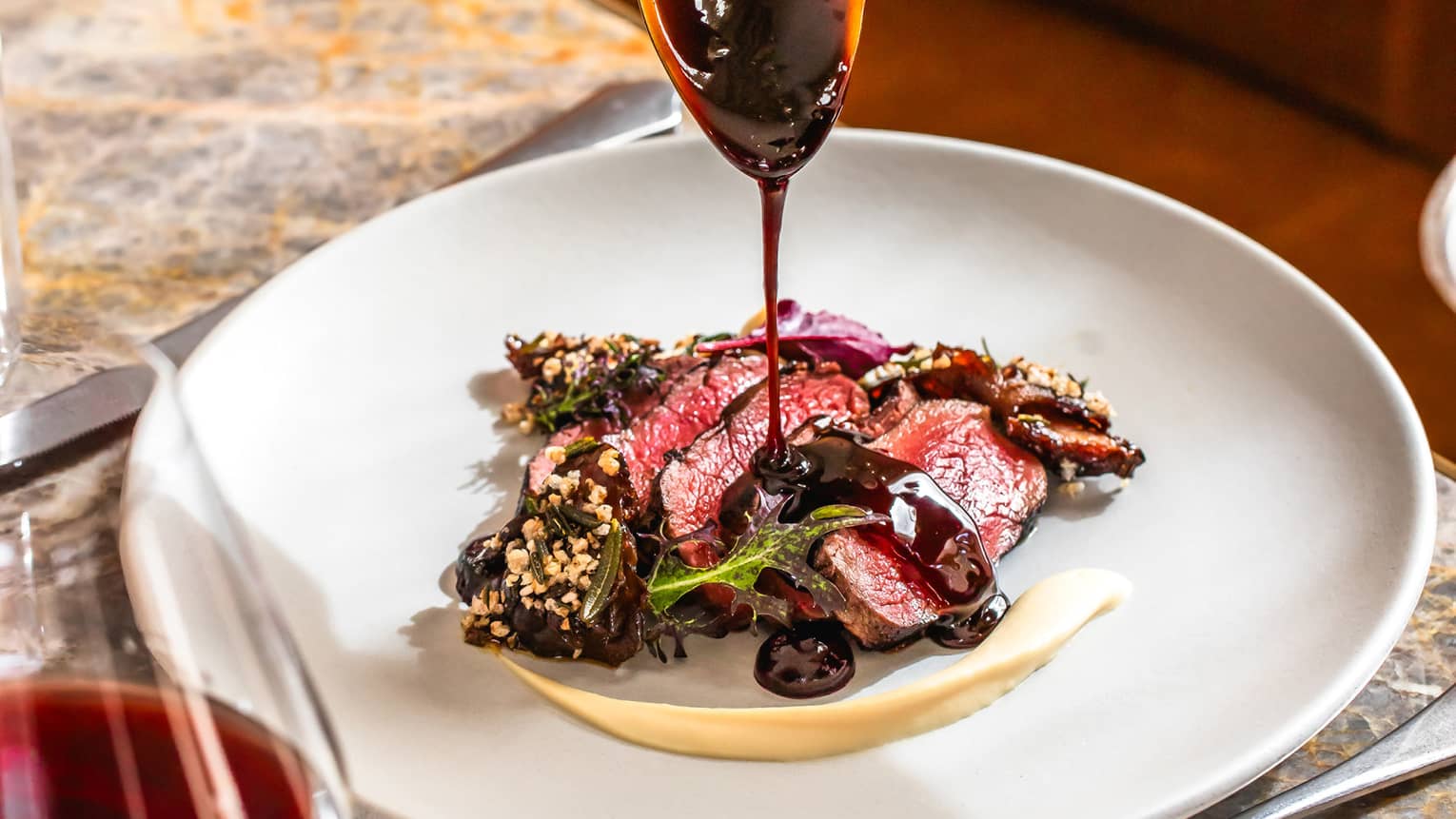 A dark sauce is being drizzled from a spoon onto food on a plate, set on a marbled tabletop. A stemmed glass of red wine is in the foreground.