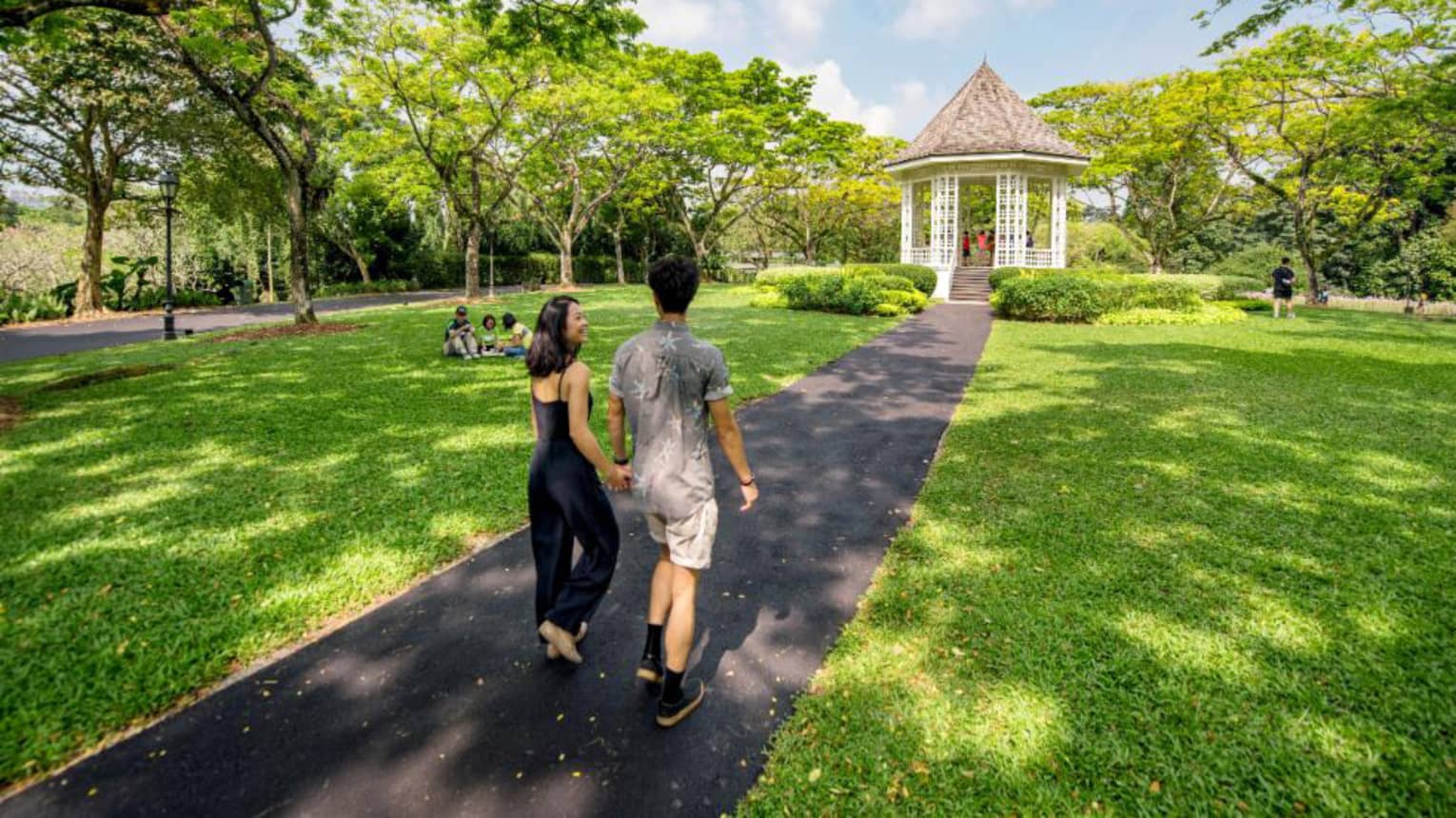 A man and woman hold hands and walk along an asphalt trail through the Singapore Botanic Gardens