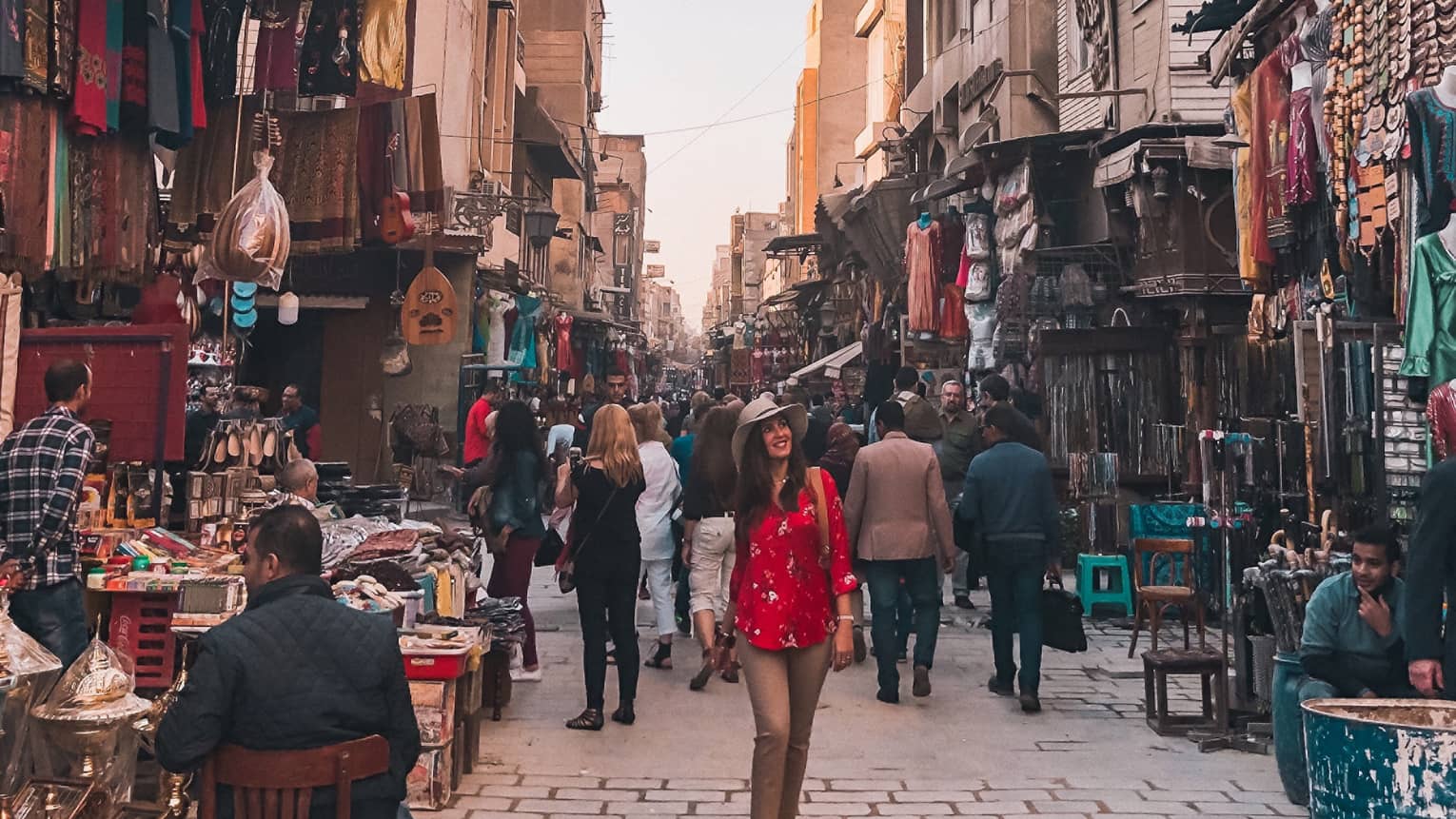 A smiling tourist strolls down a market alley, surrounded by varoius wares of the many merchants crowded between buildings.