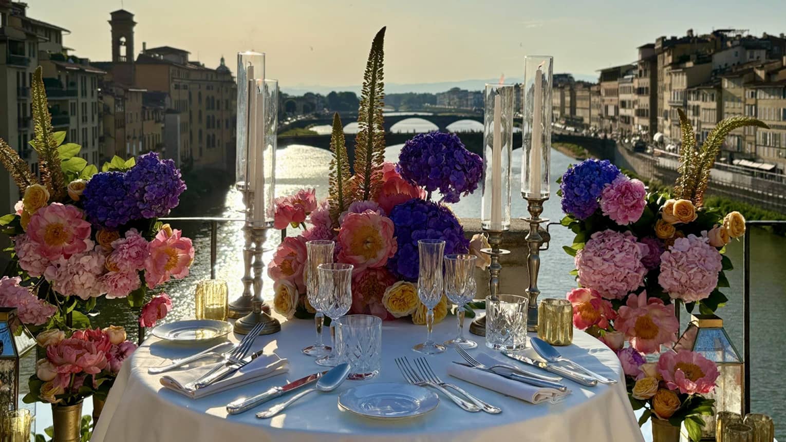 An elegantly set outdoor dining table overlooking a a river, with a sunny sky in the background.