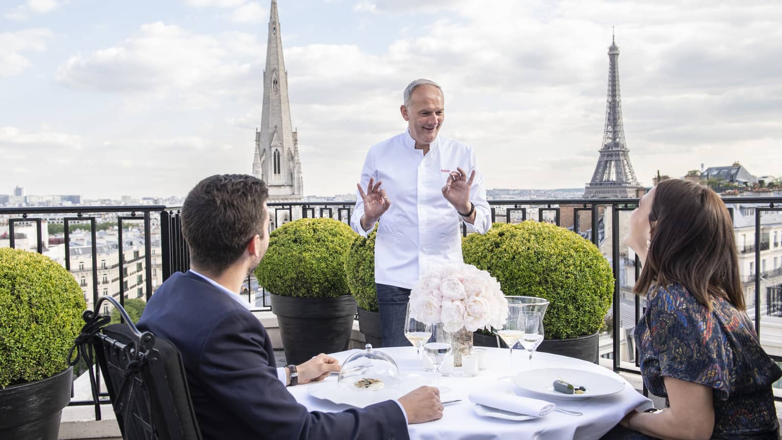 Couple seated at set dining table on penthouse terrace, chatting with Chef Christian Le Squer, Eiffel Tower in backdrop
