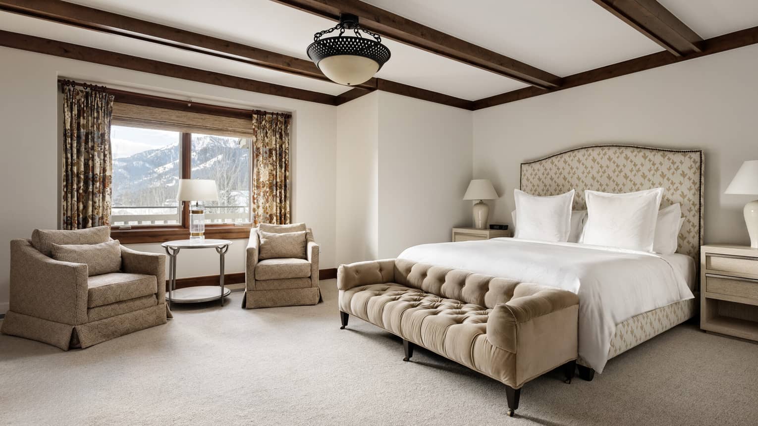 Luxury resort bedroom with a king bed, upholstered headboard, nightstands with lamps, beige tufted bench, two armchairs by the window with mountain view