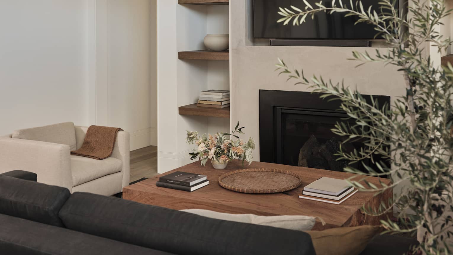 Cozy living room with beige armchair, dark gray sofa, wooden coffee table with books and flowers, built-in shelves with decor, fireplace and wall-mounted TV
