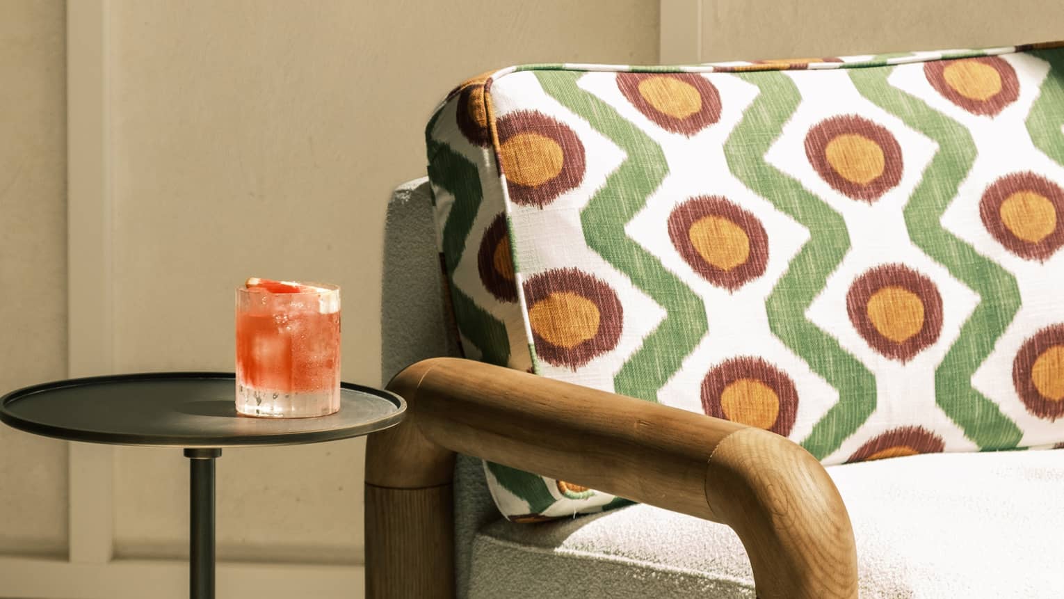 A reddish-orange cocktail in a tumbler sits on a small end table next to a couch in a partially shaded outdoor space. The couch has white seat cushions and smooth wood arms, and the back cushions are white with a geometric pattern in green, gold and red.