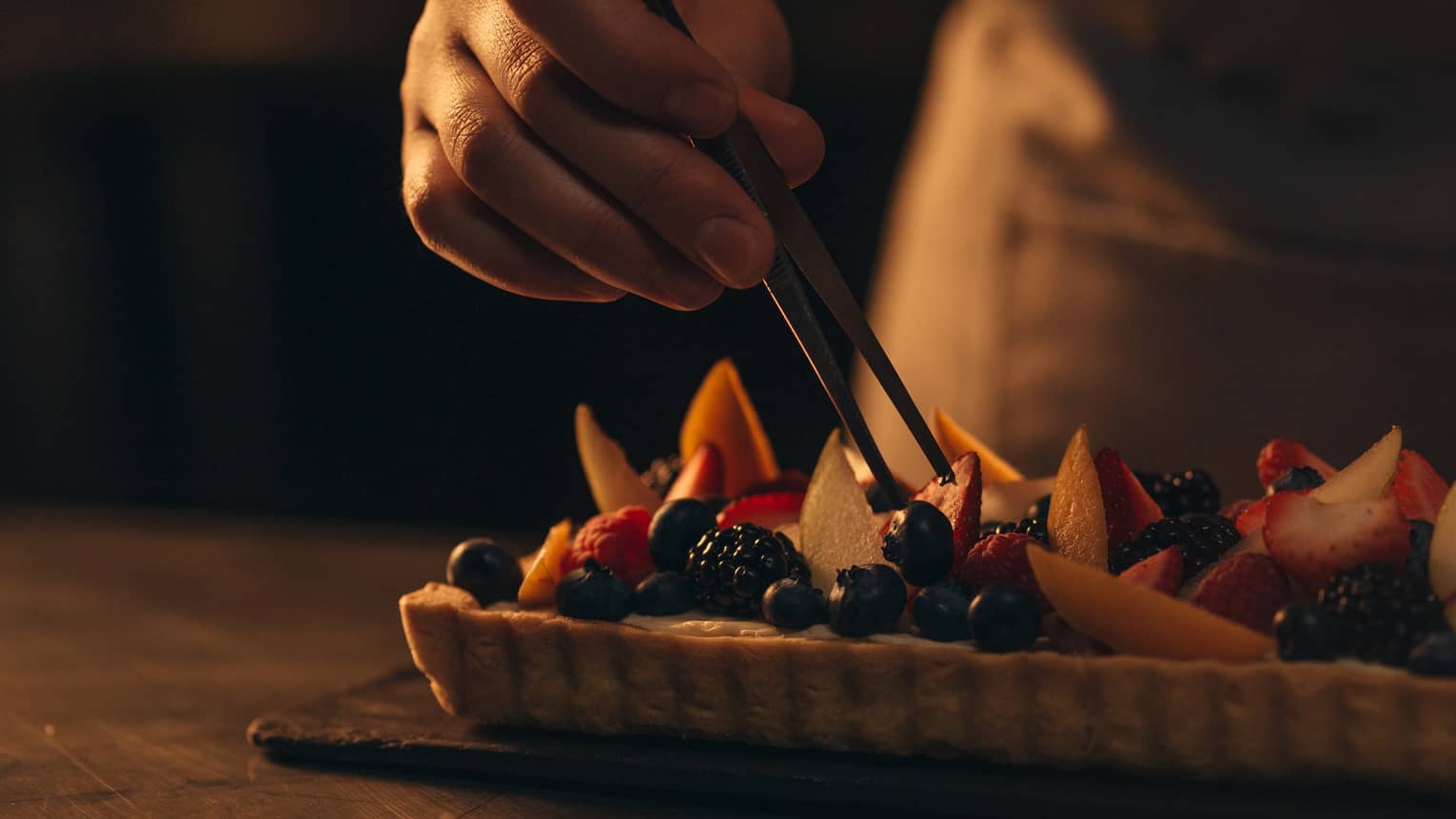 Person putting fruit slices, blueberries and blackberries onto a dessert on the table.