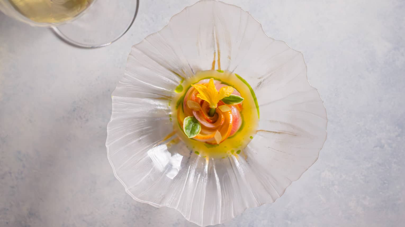 Overhead view of a colourful appetizer garnished with florals in a glass, flower-shaped dish beside a glass of white wine.