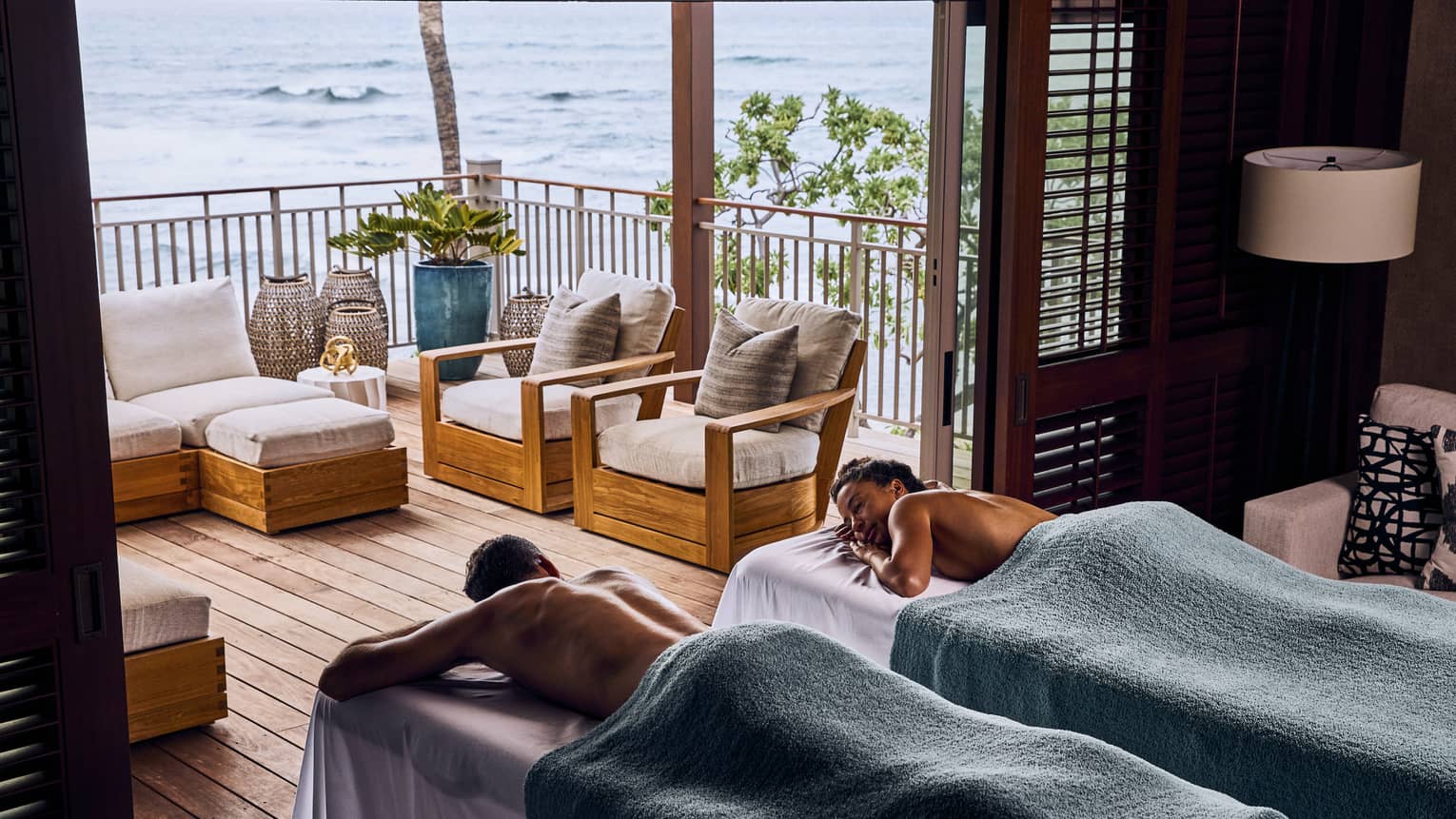 A couple covered in towels relax on massage tables in front of the open doorway of a balcony with an ocean view.