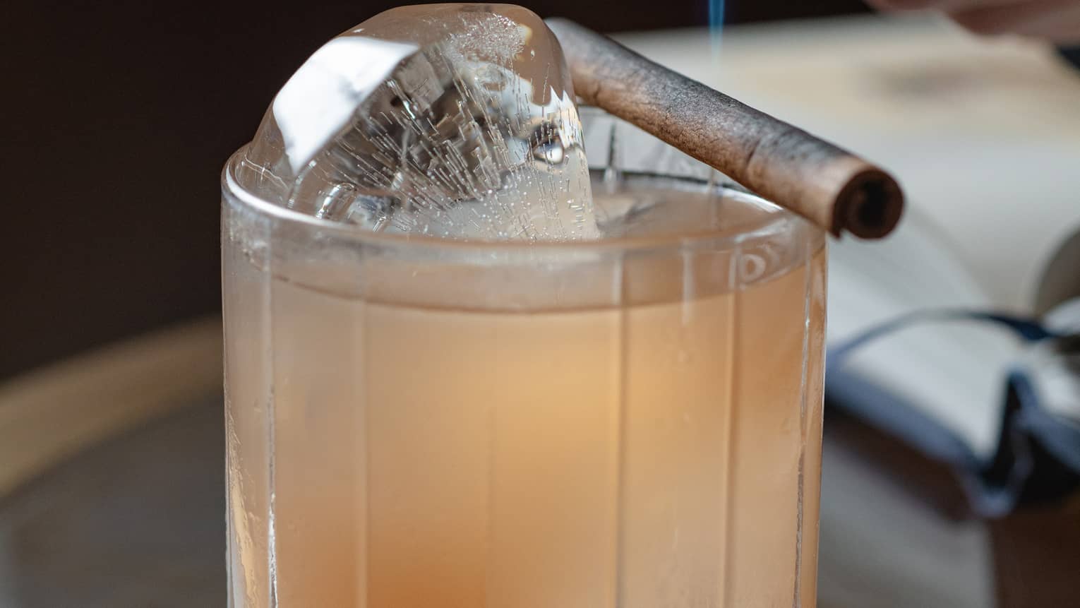 A hand holds a small blowtorch over a cinnamon stick set across the rim of a bowler glass of light amber liquid on ice.