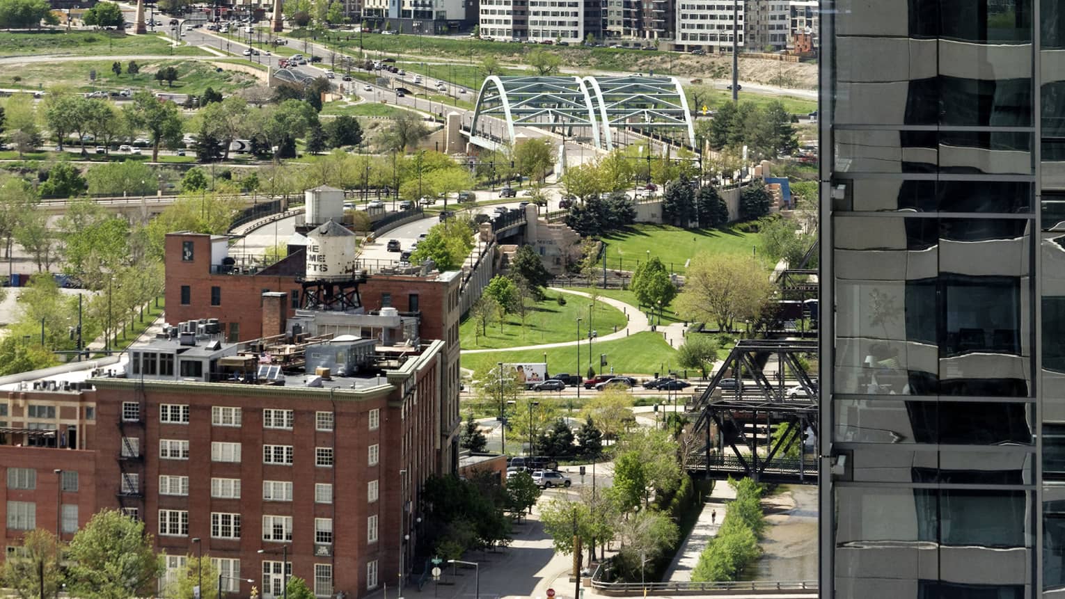 Framed on one side by a high-rise, a long aerial view of buildings and bridges amid grass and trees, misty mountains beyond.