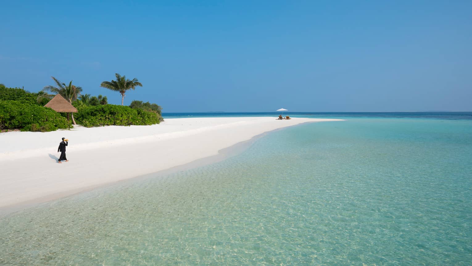 Hotel staff carries tray across long white sand beach to couple in lounge chairs by lagoon