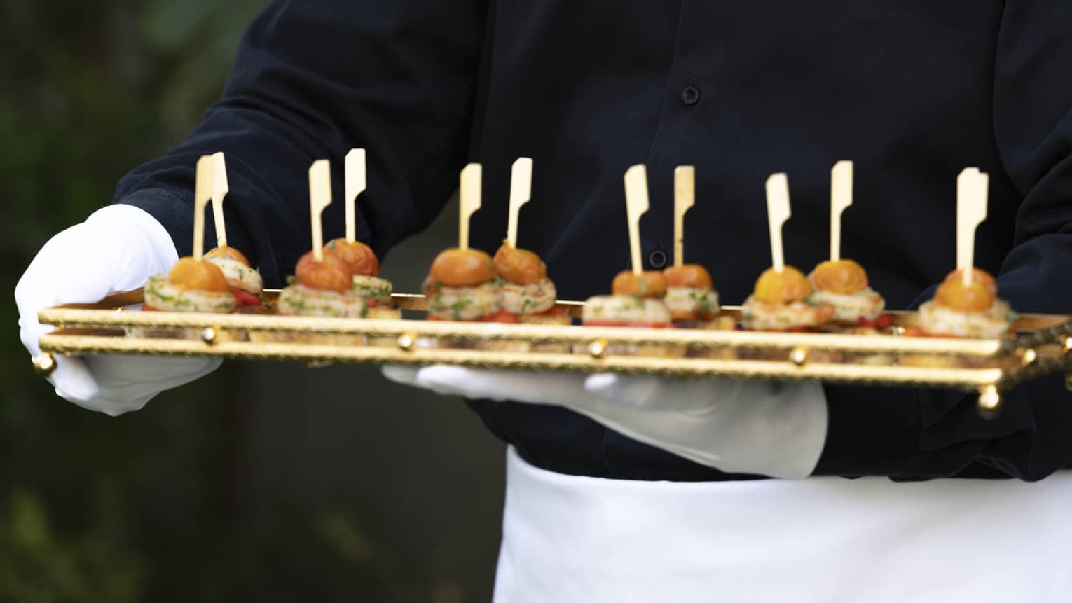 Waiter wearing black shirt, white apron and white gloves carries a tray of mini slider appetizers