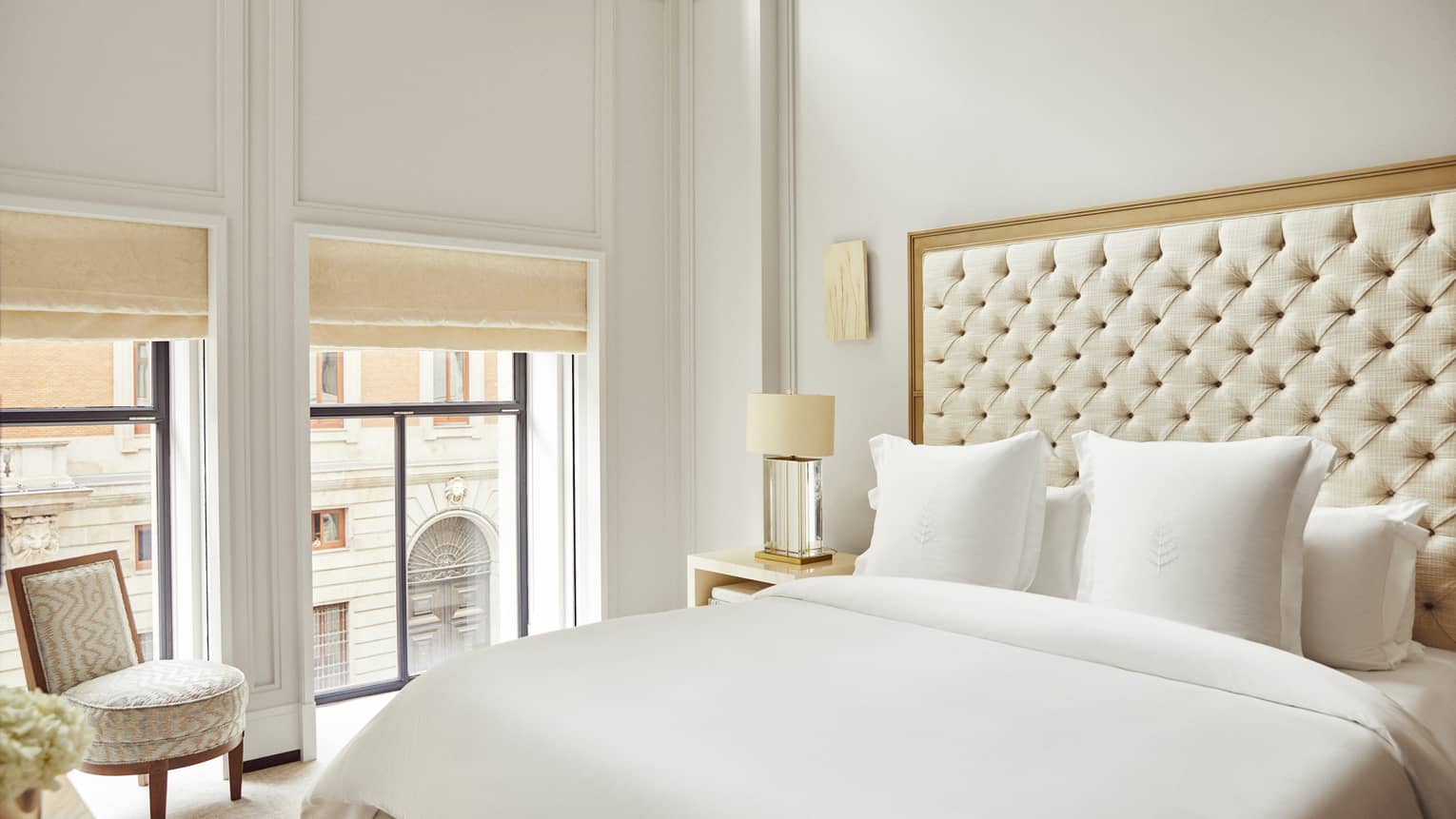 Luxury hotel suite bedroom with a king bed with tufted headboard, sleek bedside lamp, upholstered chair and windows with beige blinds