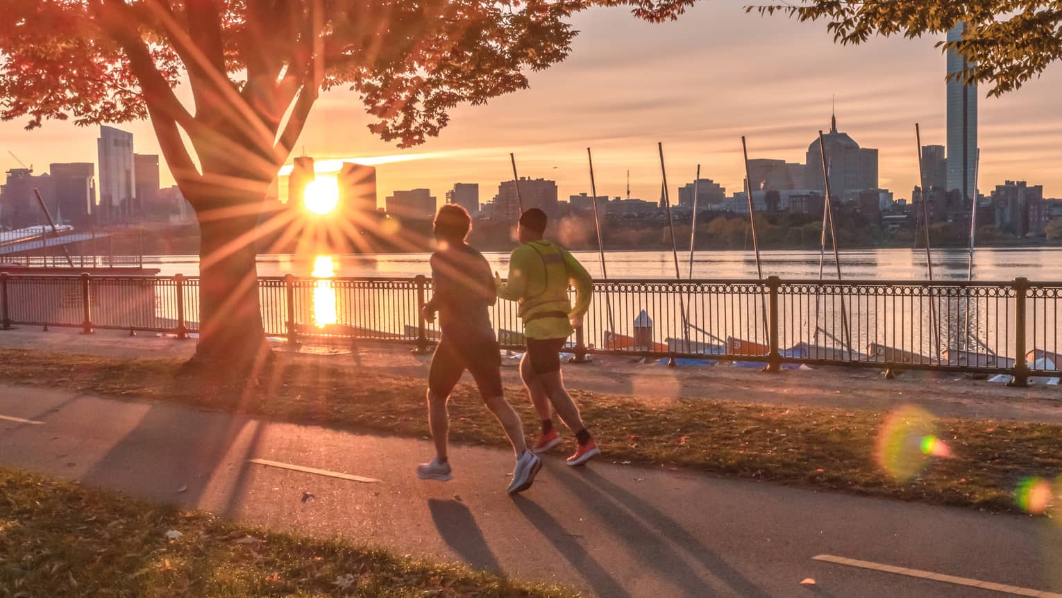 Two people jogging along a tree-lined, riverside walkway with the sun setting behind the Boston skyline